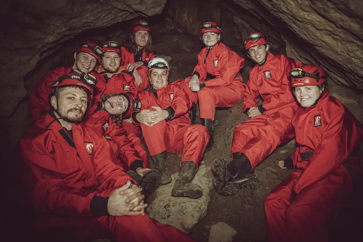 A group of trainee instructors sit together in a cave chamber, smiling, while on a caving session during an outdoor leadership and training course. They are wearing red cave suits, helmets and head torches.