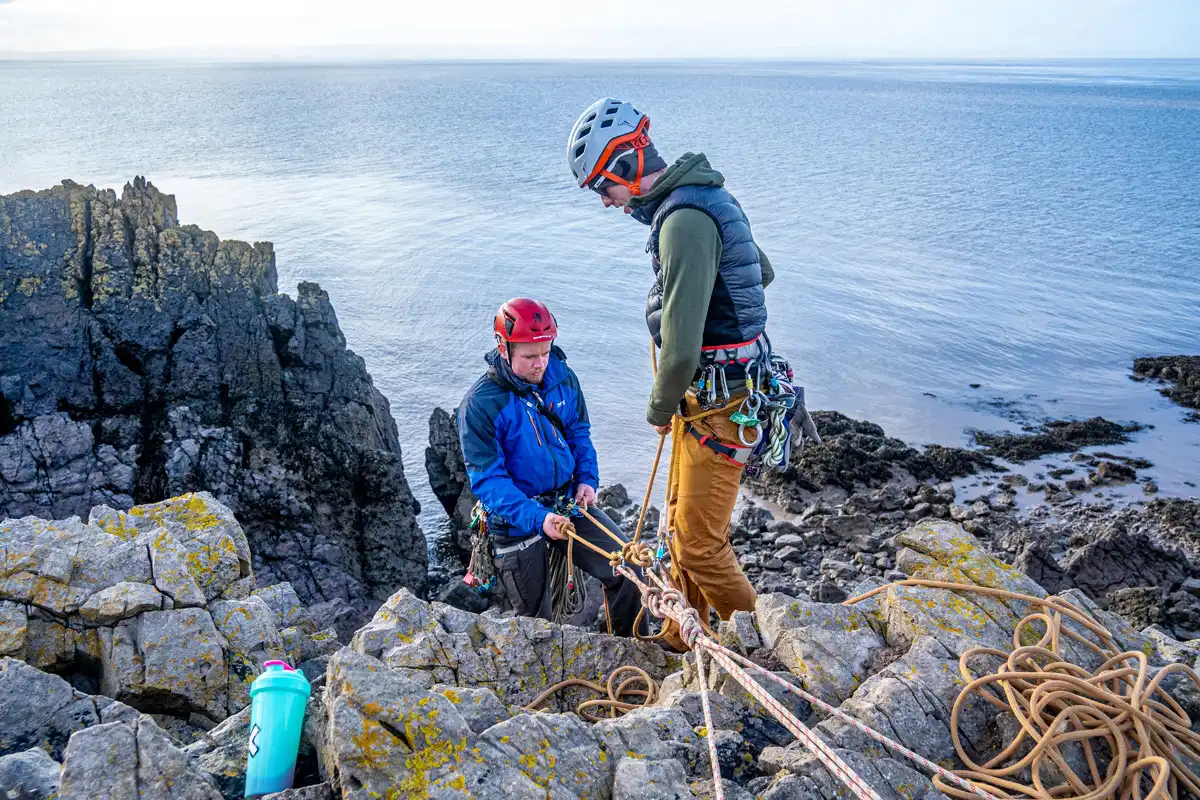 Two rock climbers are at the top of a crag, with the ocean below them spreading off into the distance. Their ropes are anchored, and both climbers are safely attached. One climber is about to lower themselves down while the other stands at the top and supervises.